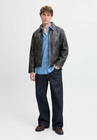 Chaqueta de cuero negra con un cuello estructurado, superpuesta a una camisa de mezclilla azul clara. Combinada con unos vaqueros oscuros sueltos y botas marrones.