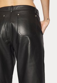 Personne portant un pantalon noir en cuir taille haute, vue de la taille jusqu'au milieu des cuisses, main reposant dans la poche droite, petit tatouage au poignet visible.
