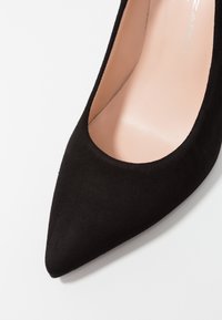 Zapato de tacón alto de ante negro con punta afilada y plantilla suave de color claro. El zapato tiene un corte profundo en los lados.