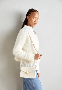 Beige läderhandväska med en strukturerad design, ett handtag upptill och en klämma i guldton. Modellen bär en fluffig vit tröja och blå jeans.