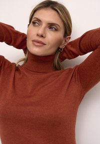 Rostfarbener Rollkragenpullover mit gerippter Textur, der enganliegende Ärmel und einen hohen Kragen aufweist, getragen mit den Händen hinter dem Kopf.