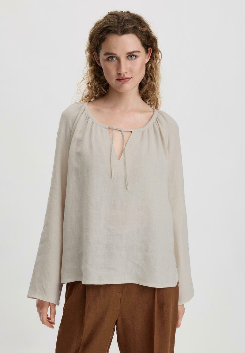Blusa in lino beige chiaro con scollatura rotonda, dettagli con nodo nella parte anteriore e maniche lunghe. Tessuto morbido e vestibilità comoda.