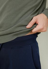 Oliwkowy sweter z ściągaczami, nałożony na granatowe dresy z elastycznym pasem i bocznymi kieszeniami.