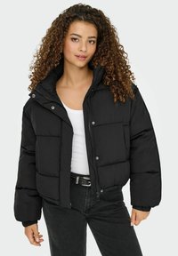 Ung kvinde med krøllet hår iført en sort dunjakke over en hvid top og sorte jeans, stående mod en ensfarvet lys baggrund.