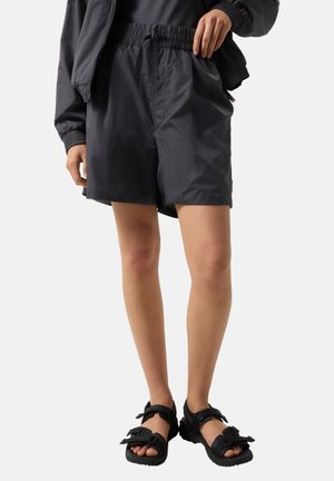 Person iført sorte elastiske taljeshorts og sorte sandaler med åbne tæer og justerbare remme, stående mod en ensfarvet baggrund.