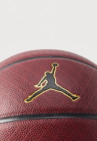 Brązowa, teksturowana piłka do koszykówki z czarno-złotym logo Jumpman przedstawiającym sylwetkę koszykarza w powietrzu z uniesioną do góry ręką.