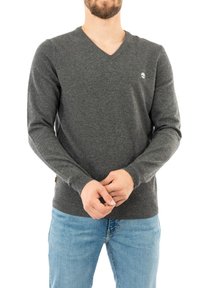 Grauer V-Ausschnitt-Pullover aus weichem Stoff, mit langen Ärmeln und einem kleinen Logo auf der linken Brust. Kombination mit hellblauen Jeans.
