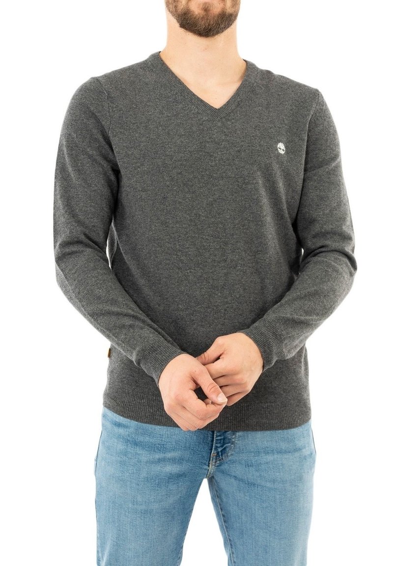 Grauer V-Ausschnitt-Pullover aus weichem Stoff, mit langen Ärmeln und einem kleinen Logo auf der linken Brust. Kombination mit hellblauen Jeans.