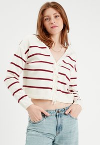 Weißer kurzer Cardigan mit maroonfarbenen Streifen, Bindegürtel vorne und langen Ärmeln, kombiniert mit hochgeschnittenen hellblauen Jeans.