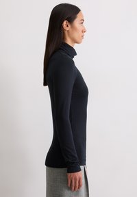 Frau, die seitlich steht, trägt einen enganliegenden schwarzen Rollkragenpullover und einen grauen Rock vor einem schlichten weißen Hintergrund.