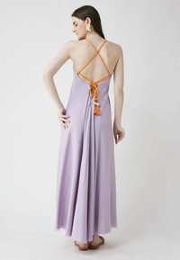 Liliowa maxi sukienka z odkrytymi plecami, z pomarańczowymi skrzyżowanymi ramiączkami i detalem w postaci frędzli w talii. Miękki, płynący materiał o lekki połysku.