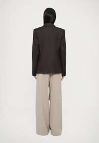 Blazer marrón oscuro con un diseño estructurado, solapas anchas y una abertura trasera única, combinado con pantalones beige de pierna ancha.