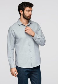 Homme barbu portant une chemise gris clair boutonnée et un jean bleu foncé, ajustant son col, debout devant un fond clair uni.