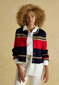 Marineblau und rot gestreifter Cardigan mit goldenen Knöpfen, der grüne und beige Akzente aufweist. Wird über einem weißen Hemd mit Knöpfen getragen. Weiche Stricktextur.