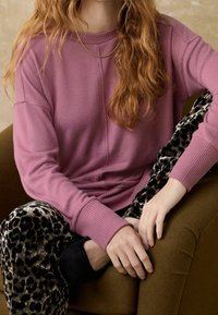 Różowy sweter z prążkowanymi mankietami, zestawiony z czarnymi spodniami w panterkę. Miękkie tekstury i wygodne dopasowanie, siedzący na brązowym fotelu.