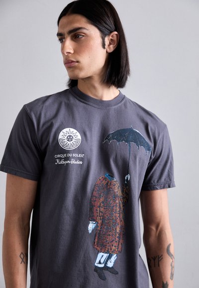 Gray t-shirt featuring a colorful graphic of a headless figure in a patterned coat holding an umbrella, with logos printed above the design.