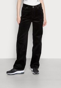 Zwarte corduroy wijde broek met zakken en metallic hardware, gestructureerd oppervlak, gedragen met zwarte sportschoenen.