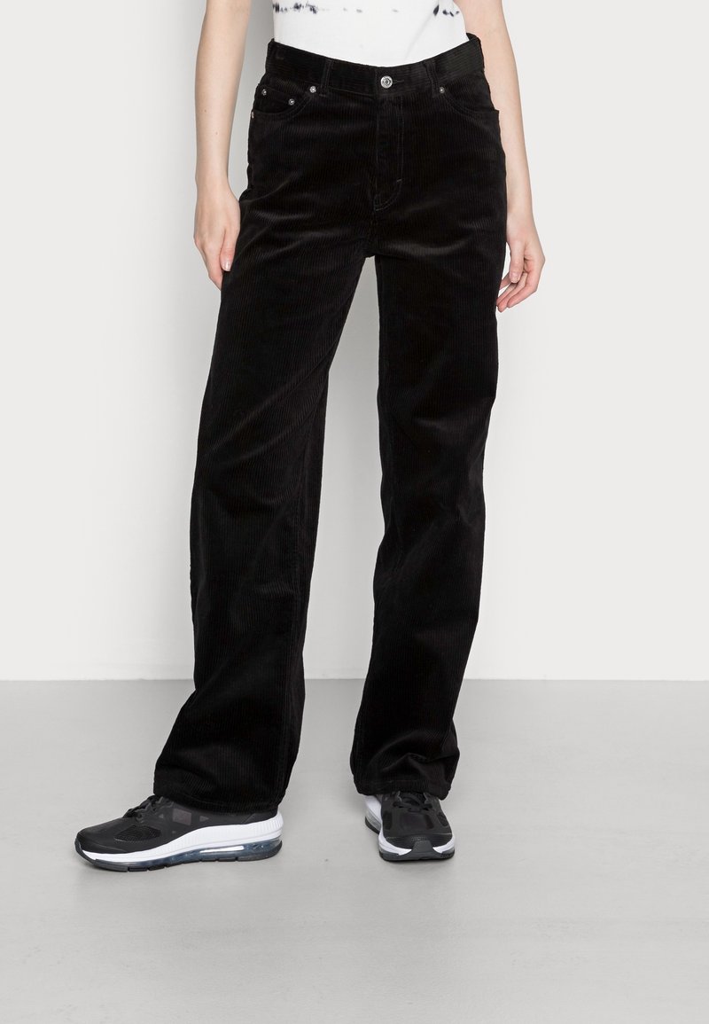 Zwarte corduroy wijde broek met zakken en metallic hardware, gestructureerd oppervlak, gedragen met zwarte sportschoenen.