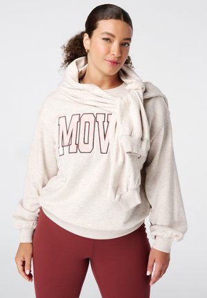 Femme aux cheveux bouclés portant un sweatshirt beige avec le texte "MOVE" et un sweatshirt assorti posé sur les épaules, assorti à un legging bordeaux.