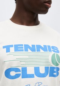 Κοντινό πλάνο ενός ατόμου που φοράει λευκό μπλουζάκι με τη φράση "TENNIS CLUB" σε μπλε γράμματα και γραφικό πράσινη μπάλα του τένις.