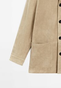 Manche de veste en daim beige avec boutons noirs visibles et grande poche avant sur fond blanc uni.