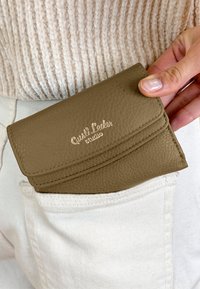 Tan leather wallet with a textured finish, featuring a flap closure and embossed logo. Positioned in a white pocket, partially visible.