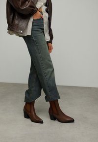 Bottes basses en cuir marron avec un bout pointu et un talon carré, portées avec un jean bleu clair retroussé et une veste texturée marron.