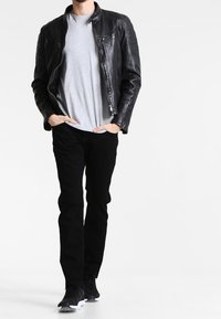 Mężczyzna idący w czarnej skórzanej kurtce, szarym t-shircie, czarnych spodniach i czarnych trampkach z białymi podeszwami na białym tle.