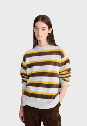 Femme aux cheveux bruns et raides portant un pull blanc avec des rayures horizontales marron et jaunes et un pantalon en velours marron, debout, une main dans la poche.