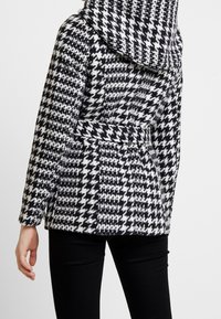 Cappotto a motivo pied-de-poule in bianco e nero, con cappuccio e cintura, caratterizzato da un tessuto testurizzato e maniche lunghe. Vista posteriore mostrata.