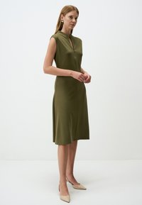 Olivegrünes ärmelloses Kleid mit leichtem V-Ausschnitt, taillierter Passform und seitlichen Schlitzen. Kombiniert mit nudefarbenen spitzen Schuhen.