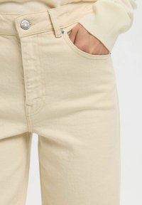 Main glissée dans la poche avant d'un jean beige, portée avec un pull en maille côtelée crème dont la manche est visible.