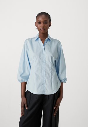 Camicia azzurro chiaro con colletto appuntito e maniche a palloncino a tre quarti. Realizzata in tessuto morbido con una texture liscia.