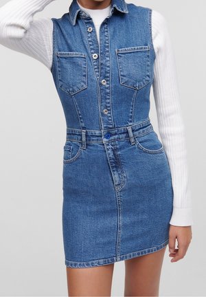 Figursyet blå denim kjole uden ærmer med knapper, forlommer og bæltestropper, båret over en hvid ribstrikket bluse med lange ærmer.