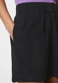 Pantalones cortos negros con una cintura elástica, bolsillos laterales y una textura suave. Costuras visibles en el dobladillo, combinados con una camiseta morada.