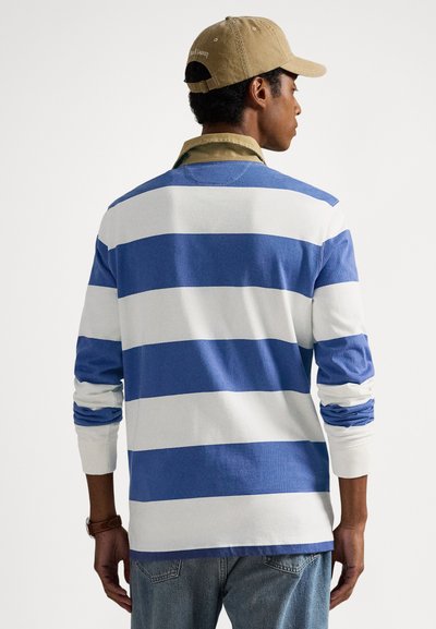 Muž v modro-bielej široko pruhovanej košeli s dlhým rukávom, s khaki golierom, modrými džínsami, hnedými hodinkami a béžovou šiltovkou, otočený chrbtom na bielom pozadí.