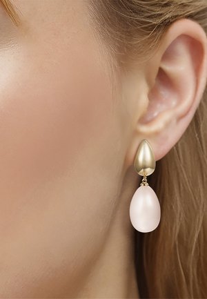 Gouden druppelsteekoorbel met een grote, gladde, witte ovale druppel, gedragen aan het oor van een vrouw met een lichte huid en blond haar.