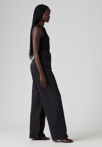 Črne široke jeans hlače z visokim pasom in klasičnim petim žepom, v kombinaciji z oprijetim črnim topom in minimalističnimi črnimi sandali.