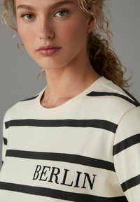 Hvid bomulds T-shirt med sorte vandrette striber, der har det broderede ord "BERLIN" i sort på forsiden. Rund halsudskæring og korte ærmer.