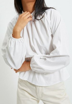 Femme portant un chemisier blanc ample à manches longues avec des poignets élastiques, associé à un pantalon beige clair, posant avec une main près du cou.