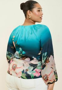 Blusa floreale con gradiente turchese e navy, maniche lunghe con polsini elasticizzati e chiusura a chiave sulla parte posteriore. Tessuto morbido e leggero.