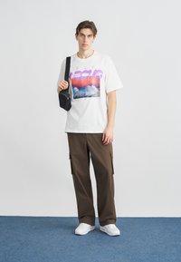 Fehér grafikás póló színes felhőmintával és a "FLY ABOVE" felirattal. Barna cargo nadrág oldalzsebekkel, valamint fehér sportcipő.