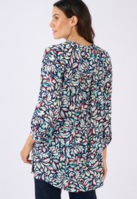 Tunica floreale con base blu navy, caratterizzata da motivi bianchi, rossi e verdi. Maniche lunghe con polsini elasticizzati, dettaglio plissettato sul retro e tessuto leggero.