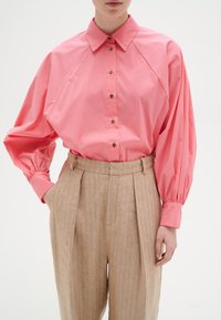 Camicia rosa oversized con colletto, chiusura con bottoni frontale e maniche voluminose. Abbinata a pantaloni a vita alta beige con righe.
