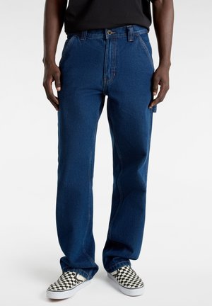 Jeans in denim blu con una linea dritta, cuciture visibili, tasche frontali e abbinati a scarpe slip-on con motivo a quadri.