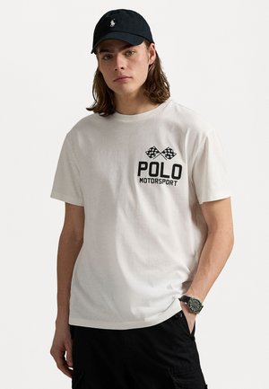 Balts kokvilnas T-krekls ar melnu "POLO MOTORSPORT" tekstu un šaha flagu grafiku, valkāts ar melnu cepuri un melniem šortiem.