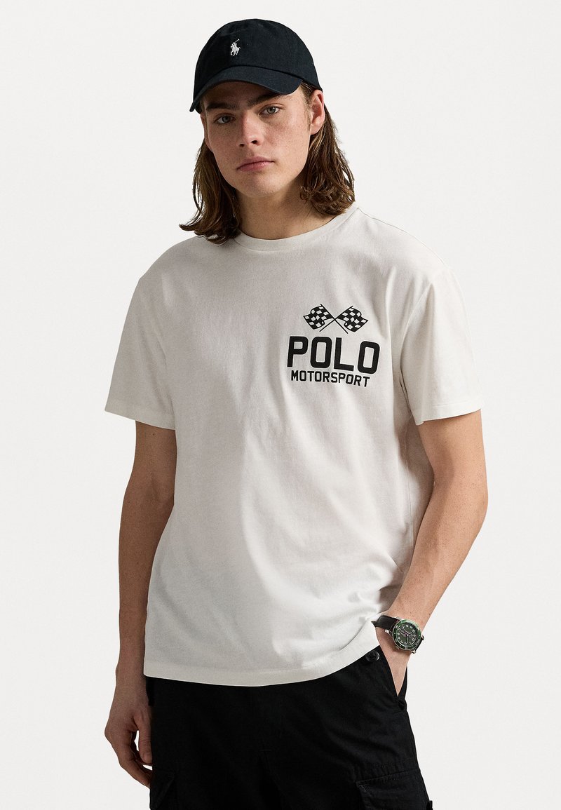 T-shirt en coton blanc avec inscription noire "POLO MOTORSPORT" et graphique de drapeau à damier, porté avec une casquette noire et un short cargo noir.