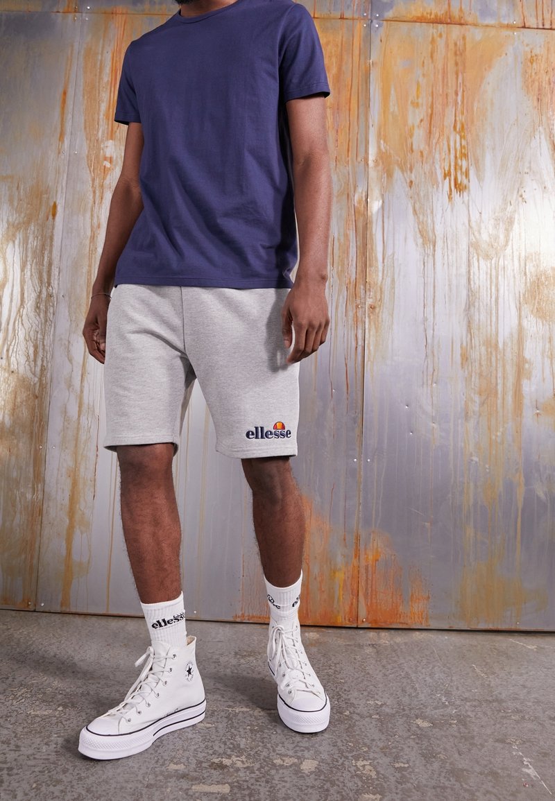 T-shirt à manches courtes bleu marine, short de survêtement gris avec logo brodé, baskets montantes blanches et chaussettes à motifs. Tenue décontractée et sportive.