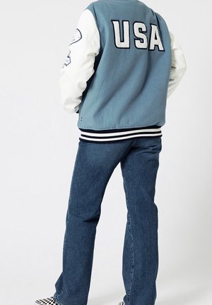 Persona que lleva una chaqueta universitaria azul y blanca con "USA" en la espalda, jeans azules y zapatos slip-on a cuadros en blanco y negro.