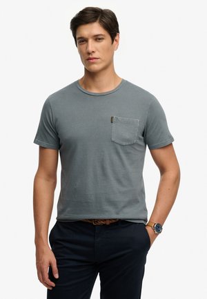 Mann mit kurzen dunklen Haaren, der ein graues kurzärmeliges T-Shirt mit Tasche, dunkle Hose, braunen geflochtenen Gürtel und eine Armbanduhr trägt.